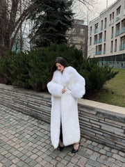 Woman in a full-length white faux fur coat, adjusting the lapels, standing on a cobblestone walkway.