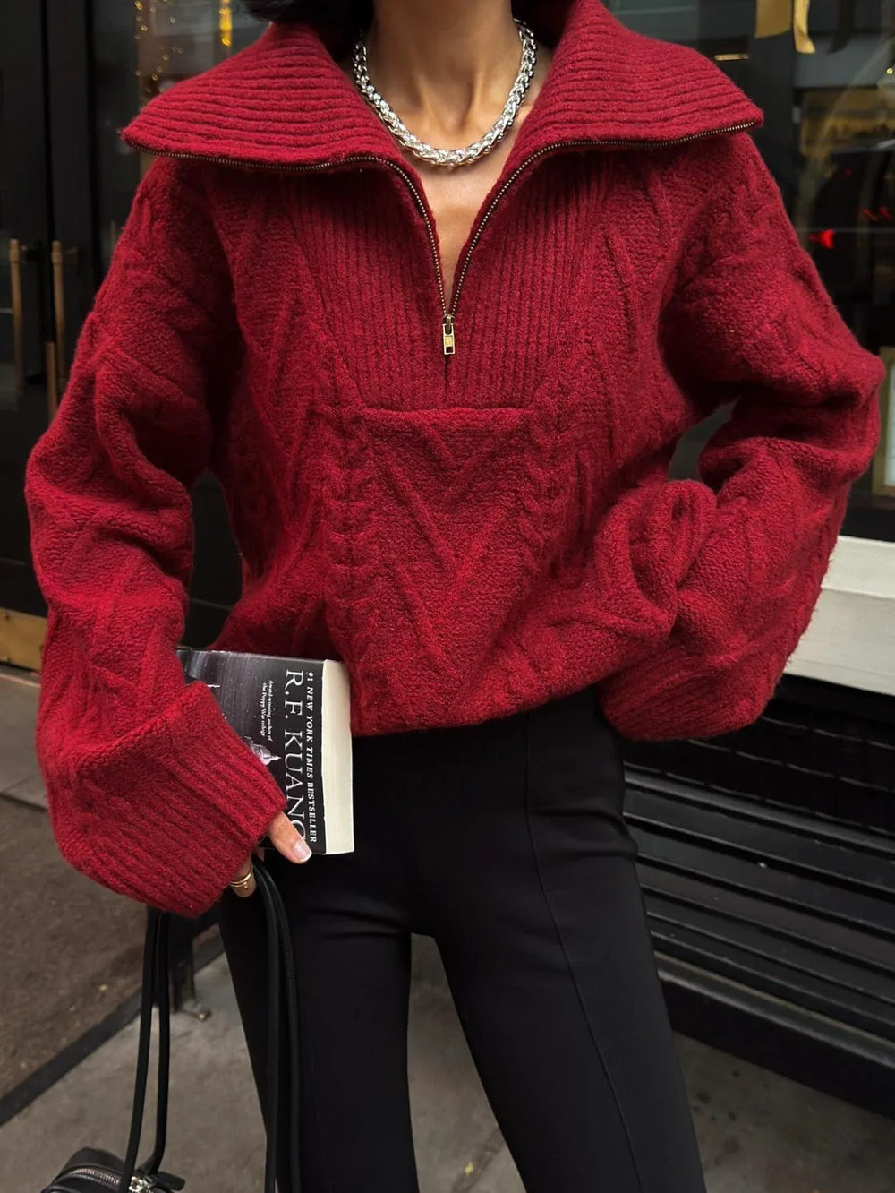 Woman wearing a red textured knit quarter zip sweater and black pants, holding a book and a handbag.