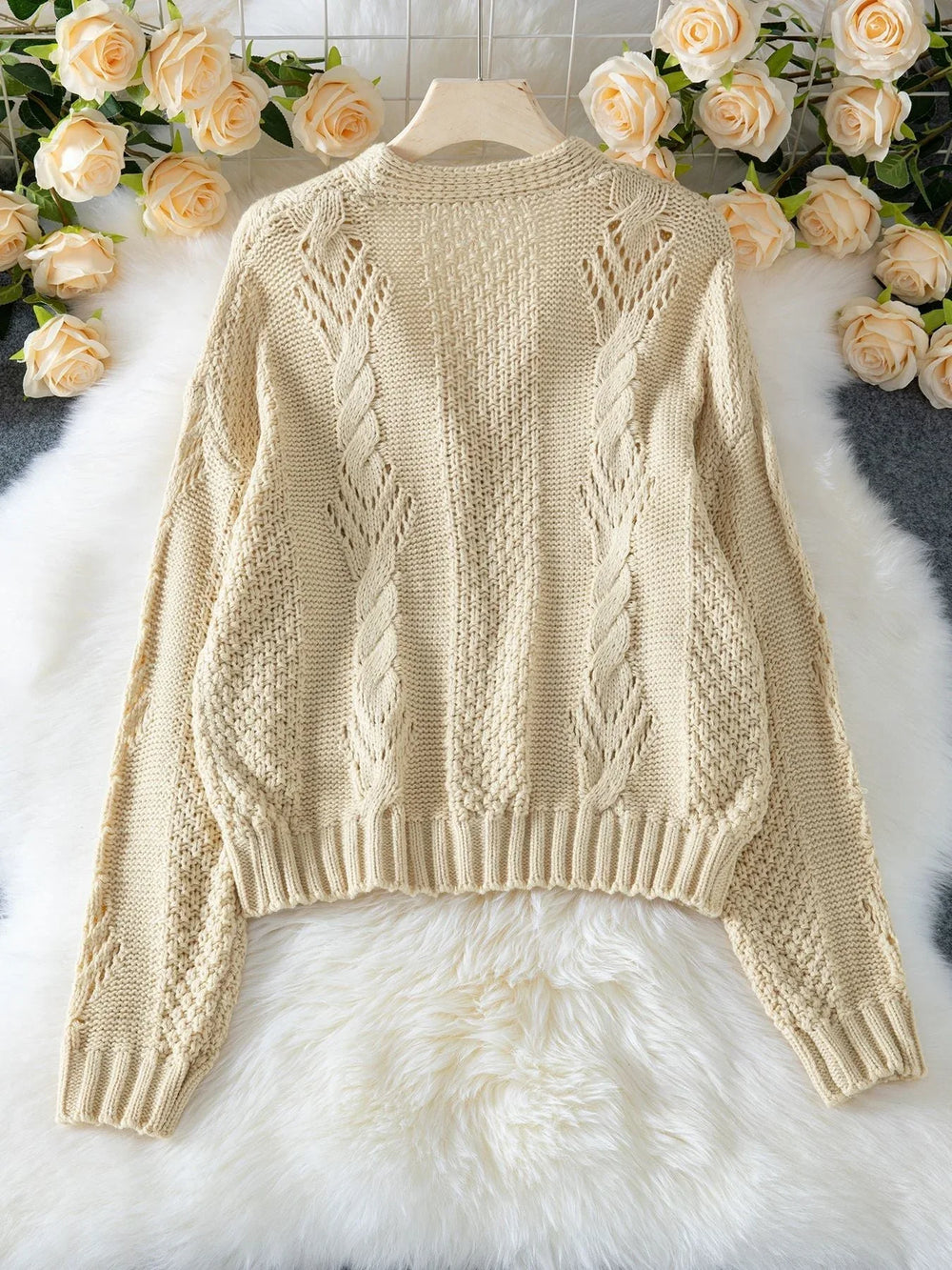 Back view of a cream-colored, open-knit, cropped cardigan with a V-neck and button-up front, laid flat on a white faux fur rug with floral accents.