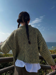 Back view of a woman wearing a green and black striped knit cardigan with colorful buttons, arms outstretched, by the ocean.