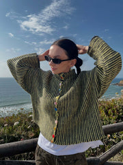 Woman wearing a green and black striped knit cardigan with colorful buttons, adjusting her hair, by the ocean.