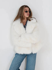 Woman wearing a white faux fur jacket over a denim shirt and blue jeans, leaning against a white wall.
