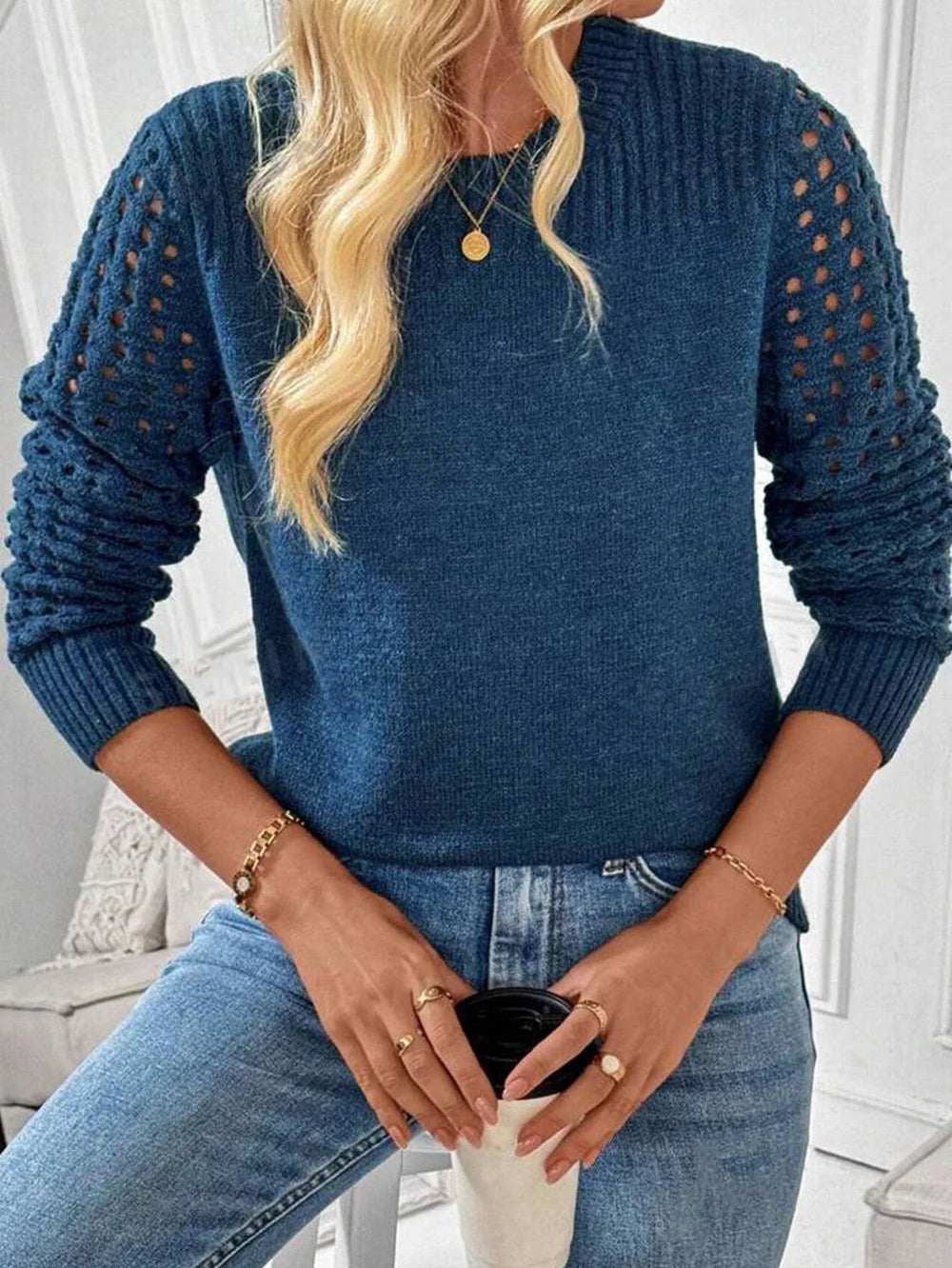 Close-up of a woman wearing a blue knit sweater with cutout sleeves and blue jeans, holding a coffee cup and wearing gold jewelry.