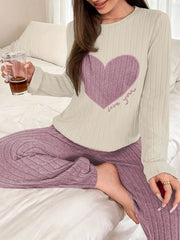Woman sitting on a bed, holding a glass with a straw, wearing a cream long-sleeve pajama top with a pink heart graphic and "love you" text, and matching ribbed purple pajama pants.
