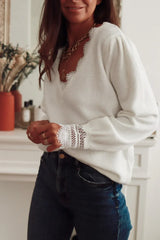 Woman wearing a white knit sweater with lace trim and blue jeans, hands clasped in front.