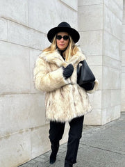 Woman in a cream faux fur coat, black hat, and black pants walking outdoors.