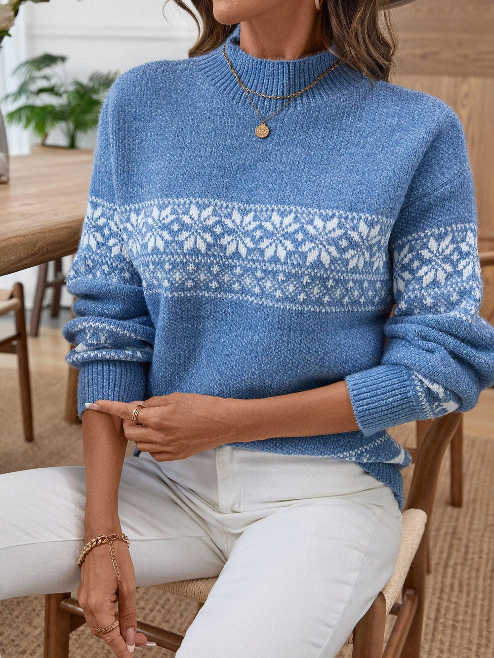 Woman sitting and wearing a blue knit sweater with a white Fair Isle snowflake and reindeer pattern, white pants.