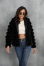 Woman wearing a black faux fur hooded jacket over a white crop top and blue jeans, with sunglasses on.