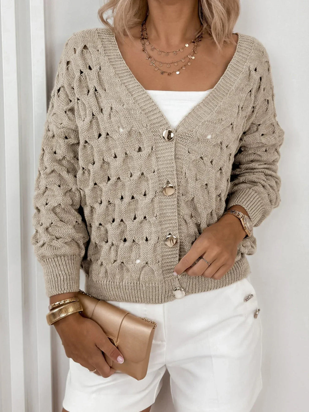 Woman wearing a beige open-knit V-neck cardigan with gold buttons, a white tube top, and white shorts.