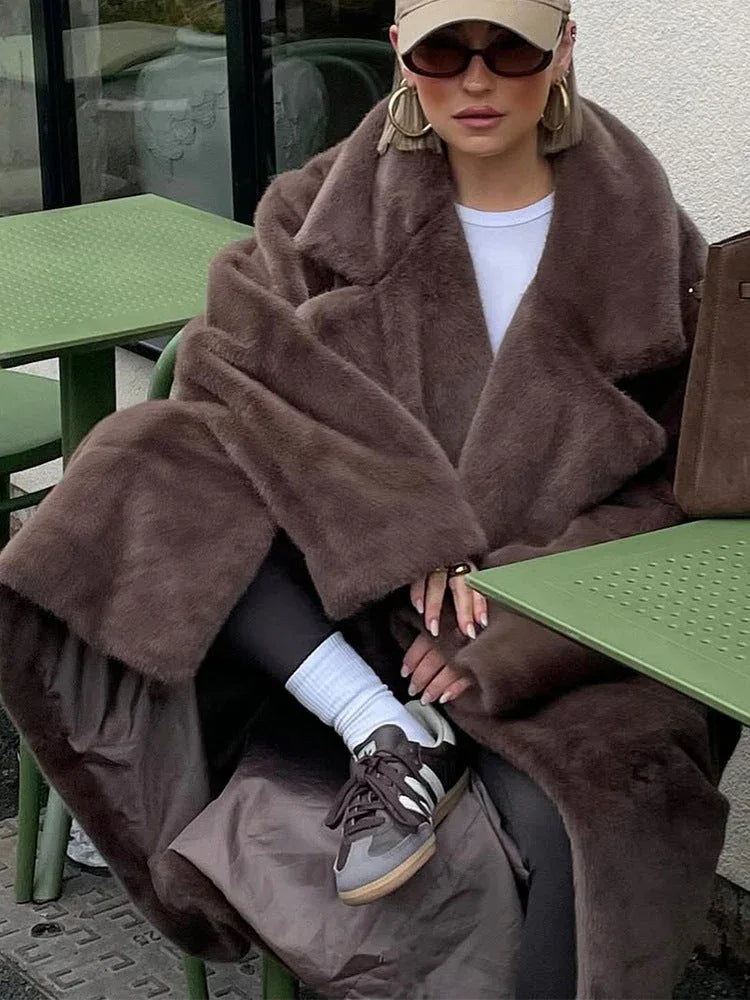 A woman is seated at an outdoor cafe, draped in a plush brown faux fur coat, with her hands resting on the table.