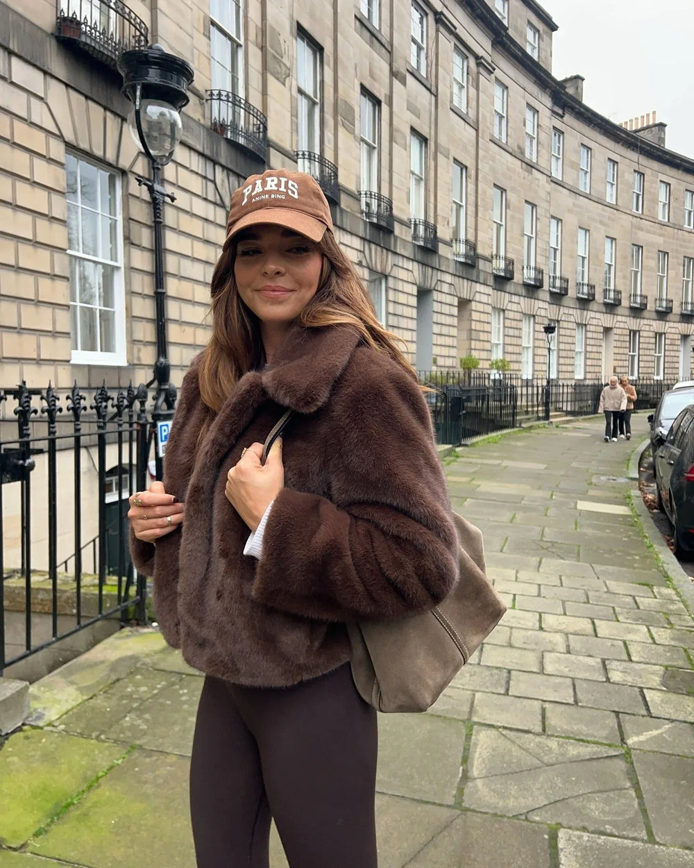 Woman wearing a brown faux fur cropped jacket, white crop top, and brown leggings, smiling at the camera.