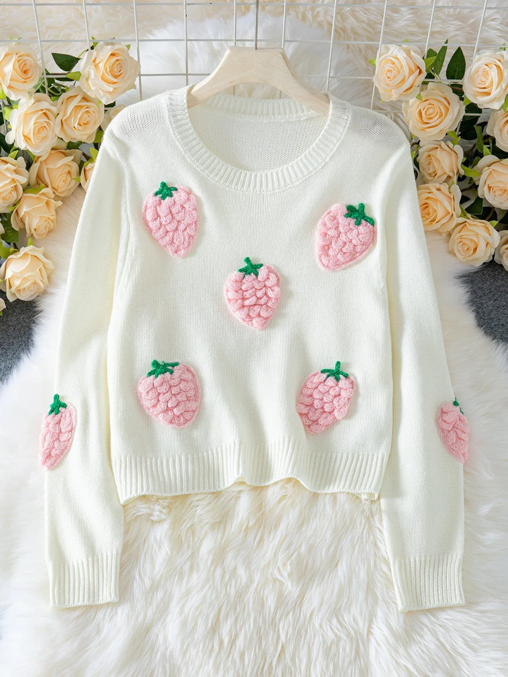 White cropped knit sweater with multiple pink strawberry appliques, displayed on a hanger with a floral background.