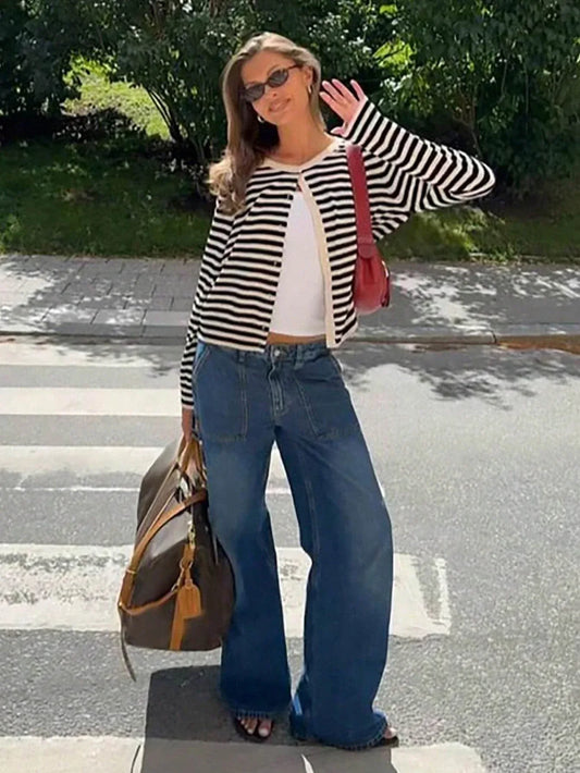 Woman wearing a black and white striped cardigan over a white tank top and wide-leg blue jeans, holding a brown duffel bag and a red shoulder bag, standing on a crosswalk.
