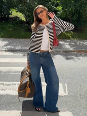 Woman wearing a black and white striped cardigan over a white tank top and wide-leg blue jeans, holding a brown duffel bag and a red shoulder bag, standing on a crosswalk.