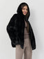 Woman wearing a black hooded faux fur jacket, brown ribbed turtleneck top, and brown wide-leg trousers, smiling and looking forward.