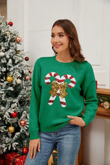 Woman wearing a green long-sleeved crew neck sweater with a sequined candy cane graphic and bow, smiling and posing in a festive Christmas setting.