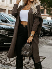 Woman wearing a long brown knit cardigan over a white crop top and black pants, holding a mini backpack.
