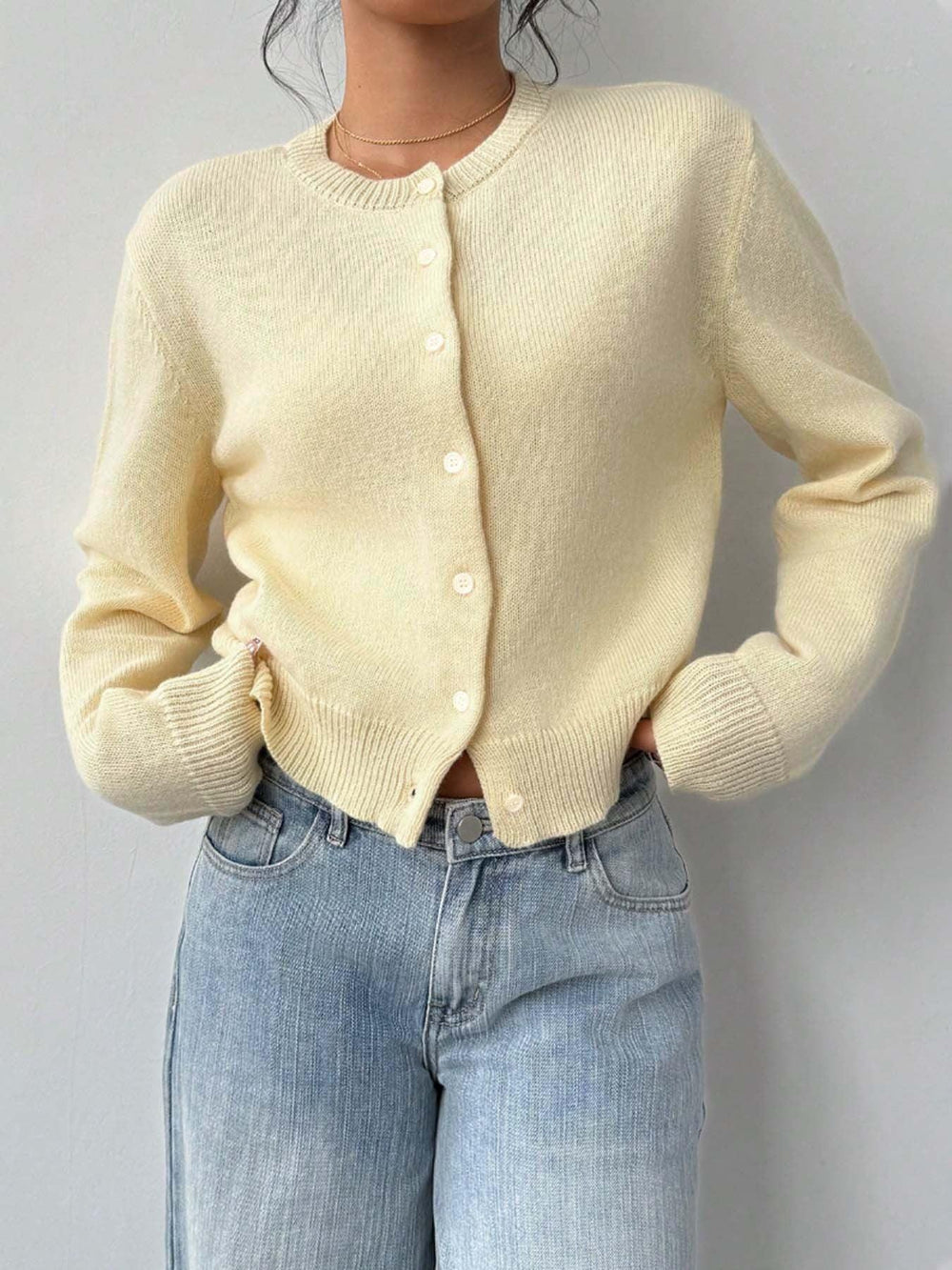 Model wearing a light yellow knit cropped cardigan with hands on hips, showing the front button closure and pairing with light-wash wide-leg jeans.