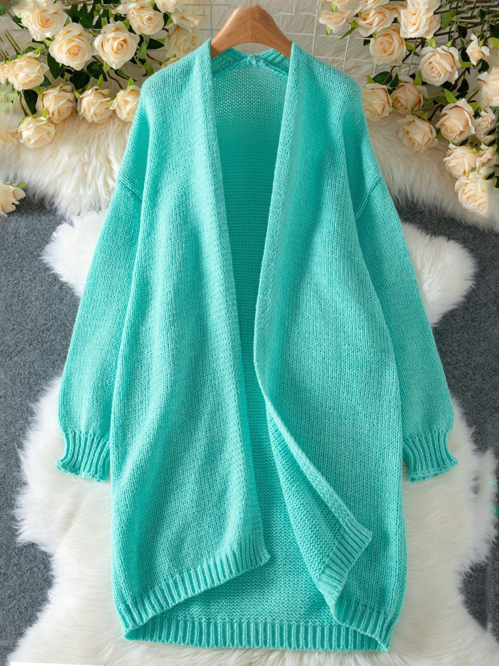 An aqua longline open-front knit cardigan laid flat on a white fluffy rug, with a floral arrangement in the background.