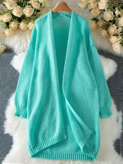 An aqua longline open-front knit cardigan laid flat on a white fluffy rug, with a floral arrangement in the background.