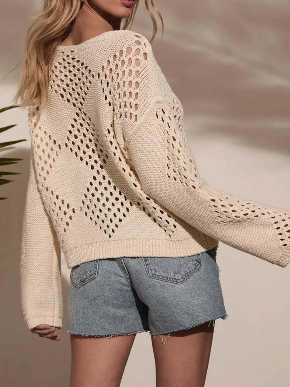 Back view of a woman wearing a cream-colored open-knit sweater with a diamond pattern, paired with distressed denim shorts.