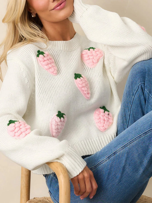 Woman wearing a white cropped knit sweater with pink strawberry appliques and blue denim jeans.