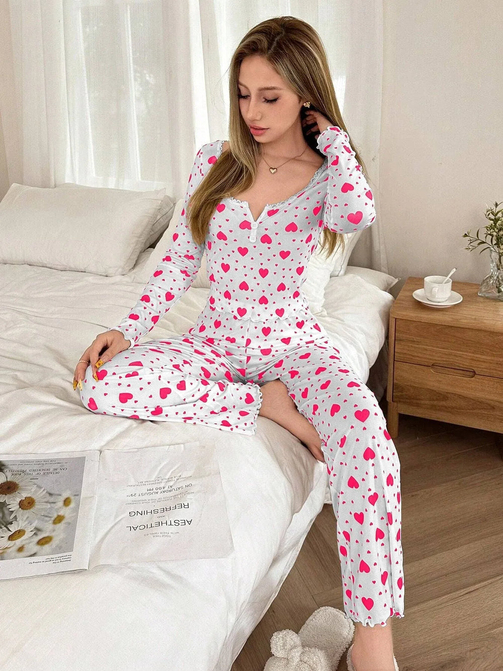 Woman sitting on a bed wearing a white long-sleeved pajama top and pants with a pink heart print, touching her hair.