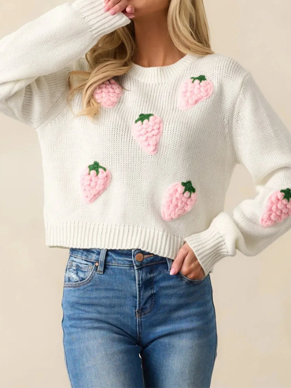 Woman in a white cropped knit sweater with pink strawberry appliques, posing with her hand on her chin.