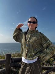 Woman wearing a green and black striped knit cardigan with colorful buttons, posing with hand raised, by the ocean.