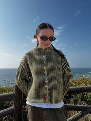 Woman wearing a green and black striped knit cardigan with colorful buttons, standing by the ocean.