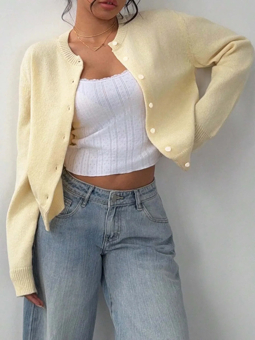 Close-up of a model wearing a light yellow knit cropped cardigan, unbuttoned to reveal a white lace-trimmed camisole, paired with light-wash wide-leg jeans.