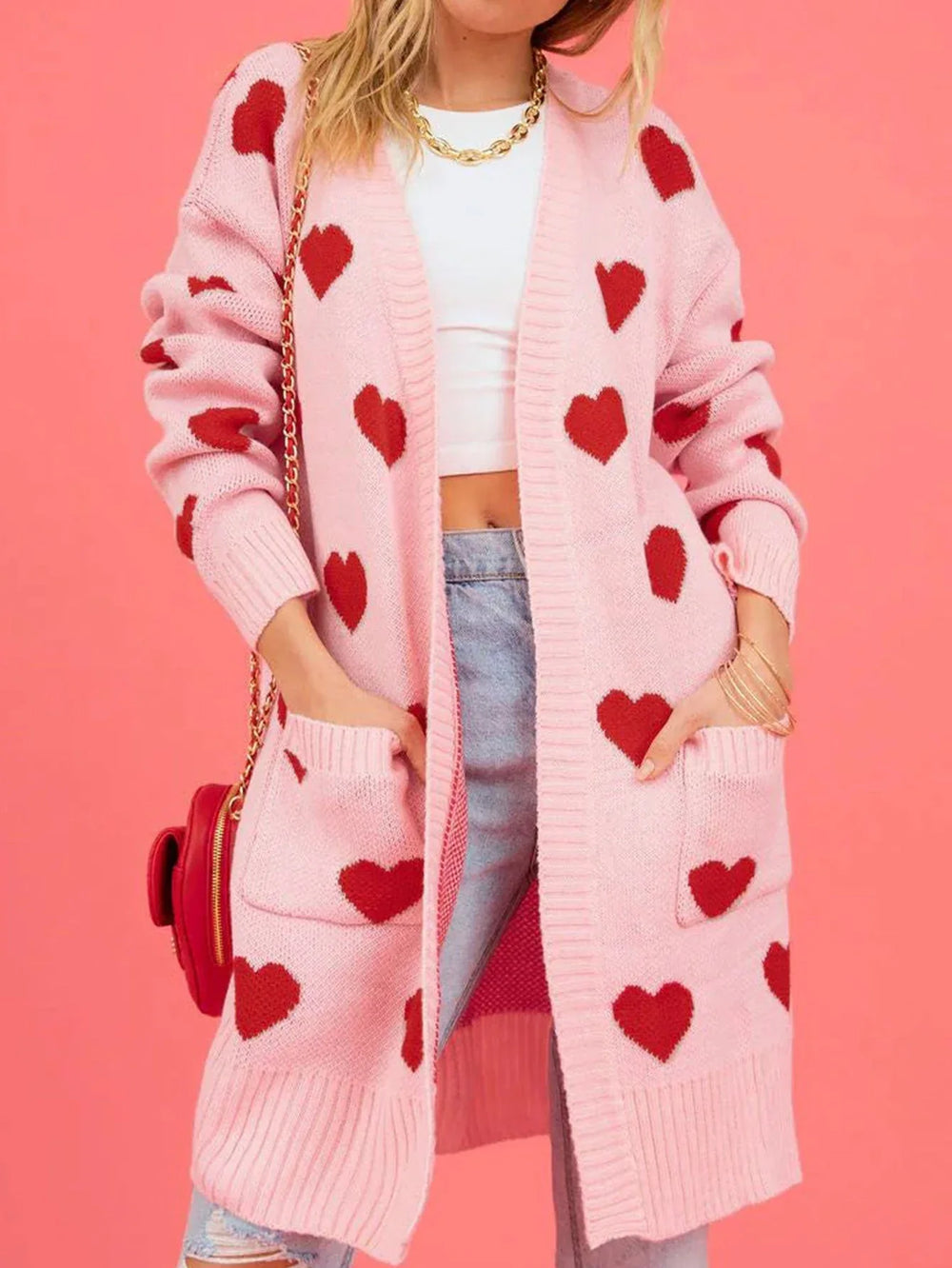 Full body shot of a woman in a pink heart cardigan, white top, and jeans, accessorized with a red handbag and gold necklace.