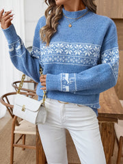 Close-up of a woman wearing a blue knit sweater with a white Fair Isle snowflake and reindeer pattern, white pants, and a white clutch bag.