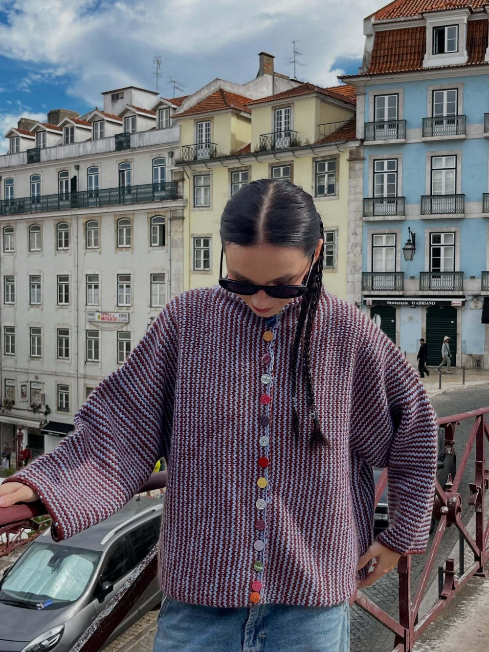 Woman wearing a burgundy and blue striped knit cardigan with colorful buttons, standing on a bridge in front of colorful buildings.