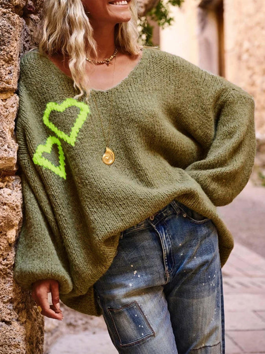 Woman wearing a green oversized knit sweater with neon yellow heart graphics on the front, paired with distressed denim jeans and a gold pendant necklace.