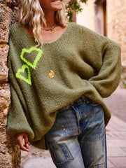 Woman wearing a green oversized knit sweater with neon yellow heart graphics on the front, paired with distressed denim jeans and a gold pendant necklace.