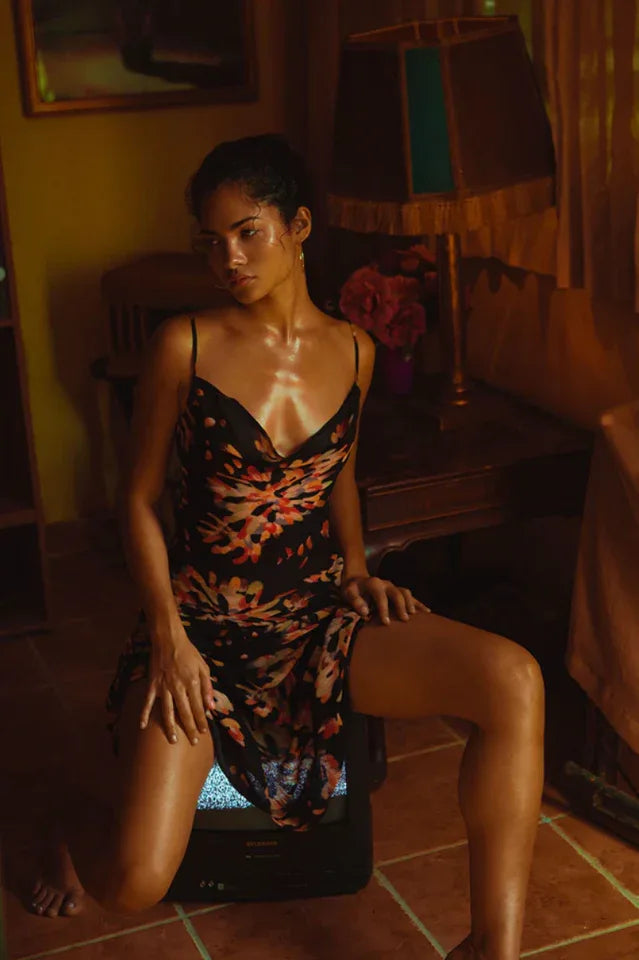 Woman wearing a black floral print maxi slip dress with spaghetti straps and a cowl neck, kneeling on a vintage television.