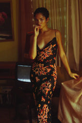 Woman wearing a black floral print maxi slip dress with spaghetti straps and a cowl neck, posing indoors with a vintage television.