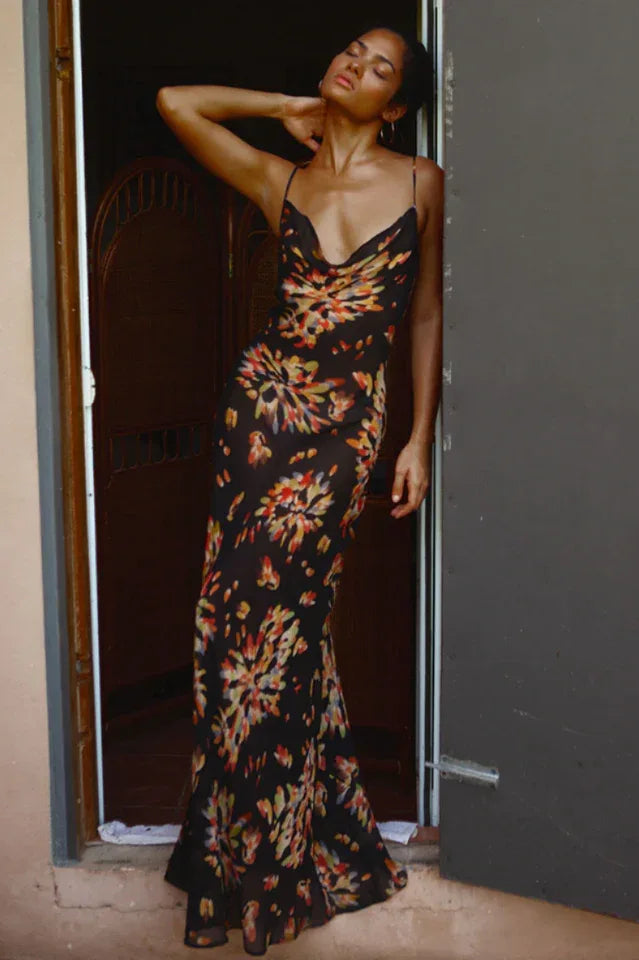 Woman wearing a black floral print maxi slip dress with spaghetti straps and a cowl neck, leaning against a doorway.