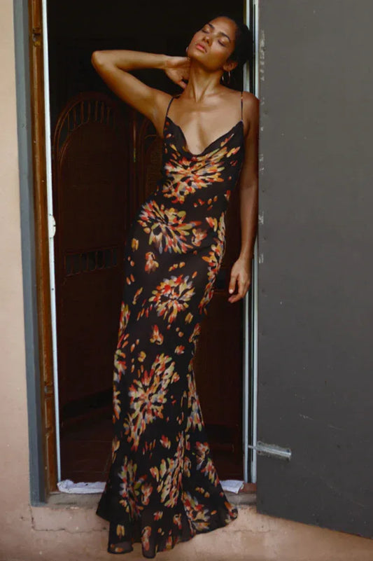 Woman wearing a black floral print maxi slip dress with spaghetti straps and a cowl neck, leaning against a doorway.