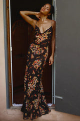 Woman wearing a black floral print maxi slip dress with spaghetti straps and a cowl neck, leaning against a doorway.