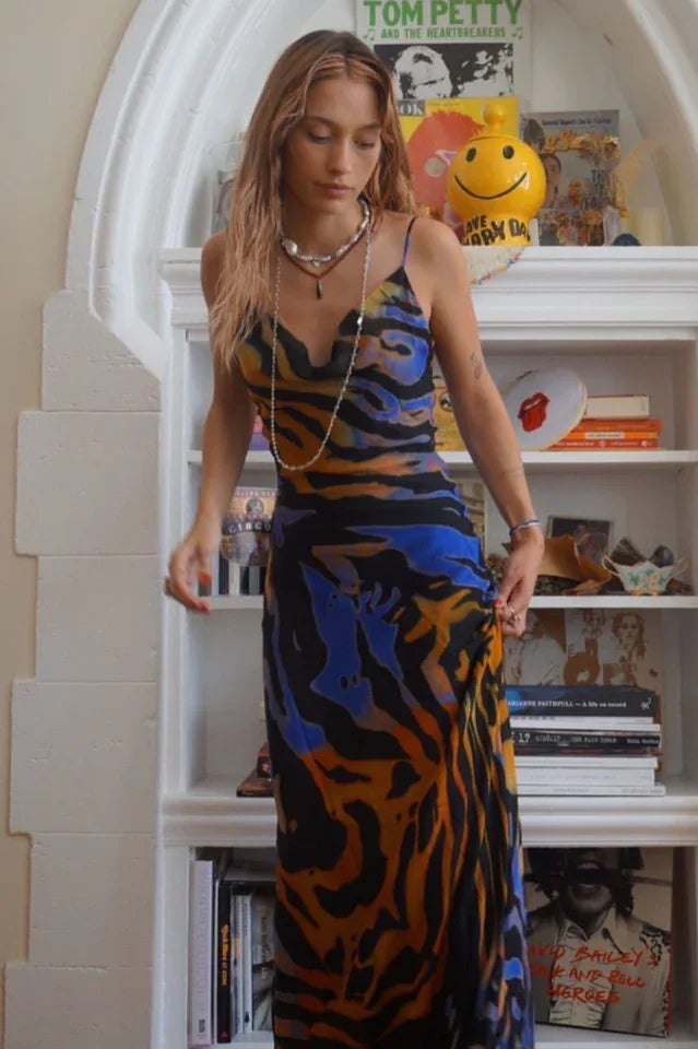Woman wearing a black, blue, orange, and yellow abstract print maxi dress with spaghetti straps and a cowl neckline, standing in front of a bookshelf.