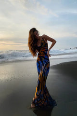 Woman wearing a black, blue, orange, and yellow abstract print maxi dress with spaghetti straps, posing on a beach with waves in the background.