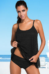 Woman wearing a black crocheted tankini top and matching high-waisted bikini bottom, posing against a beach backdrop.