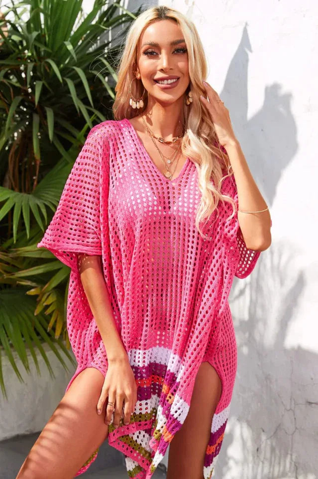 Woman wearing a bright pink crocheted cover-up with a striped hem, smiling and touching her hair.