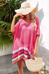Woman wearing a bright pink crocheted cover-up with a striped hem, a wide-brimmed straw hat, and sunglasses, adjusting her hat.