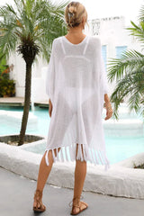 Rear view of a woman wearing a white sheer knit poncho with fringe detail over a white bikini, standing by a pool with palm trees.