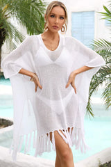Woman wearing a white sheer knit poncho with fringe detail over a white bikini, standing by a pool with palm trees.
