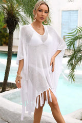 Woman wearing a white sheer knit poncho with fringe detail over a white bikini, posing by a pool with palm trees.