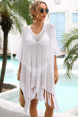 Woman wearing a white sheer knit poncho with fringe detail over a white bikini and sunglasses, standing by a pool with palm trees.